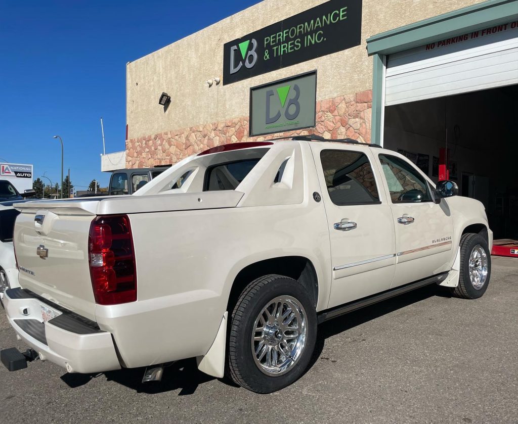 CreamÃ¢â‚¬â€˜coloured Chevrolet Avalanche with chrome aftermarket wheels and custom tonneau cover, parked outside DV8 Performance & Tires Inc. automotive shop under a clear blue sky.
