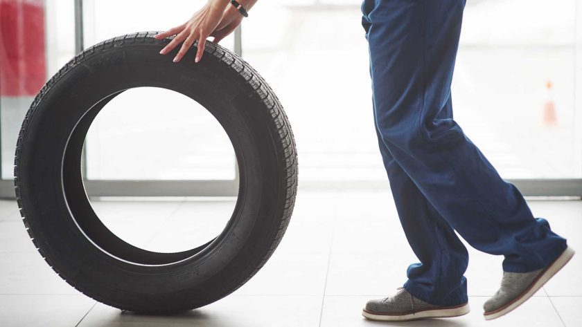 Technician rolling a black replacement tire across the tiled floor of a tire and automotive mechanic shop, showing the tireÃ¢â‚¬â„¢s tread pattern and the mechanicÃ¢â‚¬â„¢s blue work pants and gray shoes.