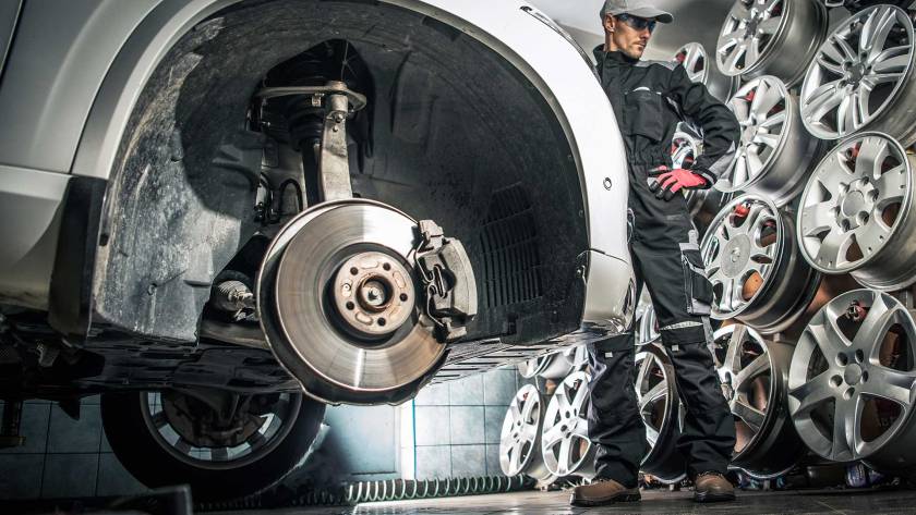 Auto technician standing beside a lifted car with the wheel removed, inspecting the brake rotor and suspension system in a tire and wheel service shop.