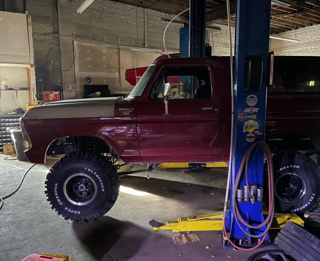 Classic red Ford Bronco lifted on a hydraulic hoist inside an auto repair shop for maintenance or restoration work.