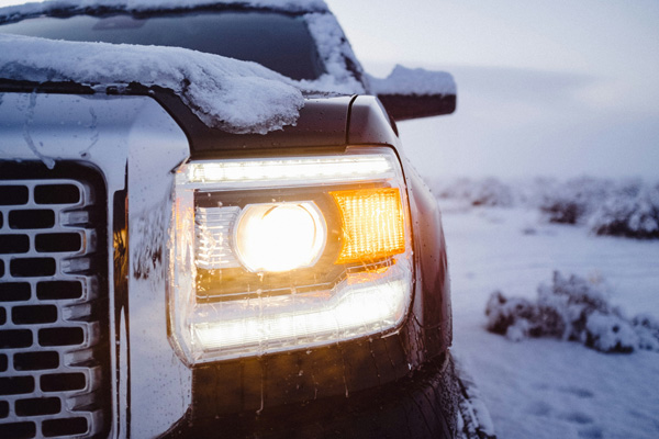 Front headlight of a truck covered in snow with lights on, parked outdoors in a snowy winter landscape.